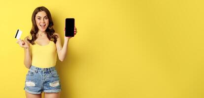 Enthusiastic modern woman showing smartphone screen, holding credit card and smiling, recommending mobile app, standing over yellow background photo