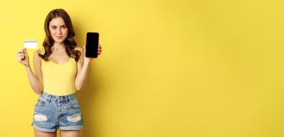 Young beautiful woman model showing smartphone empty phone screen and credit card, ready for summer, wearing tank top and shorts, standing over yellow background photo