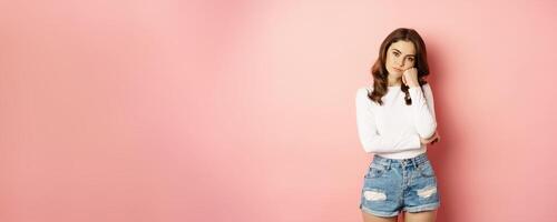 Image of sad and bored, reluctant young woman looking at smth boring, feeling lonely, leaning head on fist with indifferent face expression, standing over pink background photo