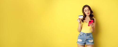 Online shopping. Stylish brunette woman holding smartphone and credit card, paying in app, using mobile phone application, buying smth, standing over yellow background photo