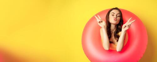 Portrait of attractive modern girl inside trendy pink beach swimming ring, showing peace v-sign and smiling happy, standing over yellow background photo