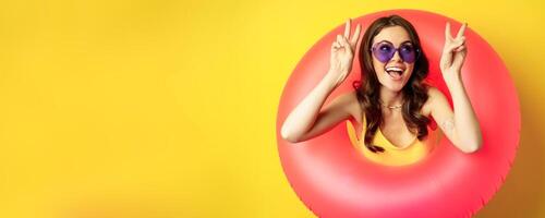 Portrait of happy stylish girl in sunglasses, inside beach swimming ring, showing peace sign, laughing and smiling, standing over yellow background photo