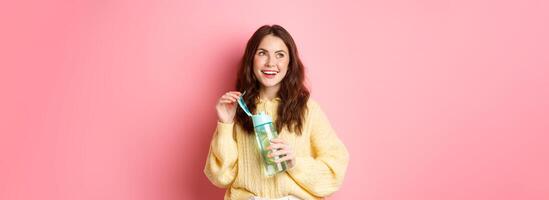 Beautiful glamour girl looking thoughtful at upper left corner copy space, reading your promotional text, drinking healthy drink from water bottle, pink background photo
