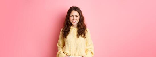 Portrait of good-looking young woman in sweater, holding hands in pockets, smiling and staring determined at camera, standing against pink background photo