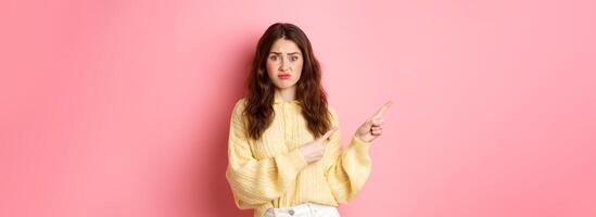 Sad and gloomy brunette girl complaining on bad promotional text, pointing fingers aside to left, showing banner with disappointed expression, standing over pink background photo