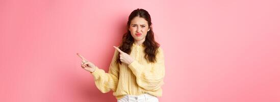 Skeptical and displeased young woman frowning, grimacing and pointing fingers right at copyspace, express dislike and disappointment, standing against pink background photo