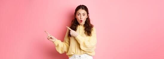 Portrait of surprised brunette girl gasping impressed, pointing fingers right at copy space aside, showing way to logo, standing over pink background photo