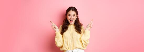 Image of cheerful good-looking female model pointing fingers aside, showing two choices sidewas, left and right logo, standing over pink background photo