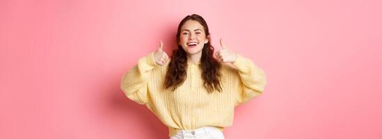 Very good, well done. Smiling girl being supportive, laughing and showing thumbs up in approval, like awesome idea, praise you, standing against pink background photo