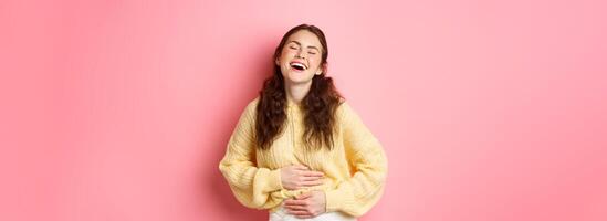 Image of carefree beautiful girl laughing out loud, touching her belly while chuckling over funny comedy, hilarious joke, standing against pink background photo