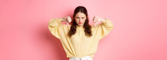 Angry and displeased young girl frowning, showing thumbs down at something bad, express dislike, leave negative feedback, standing upset against pink background photo