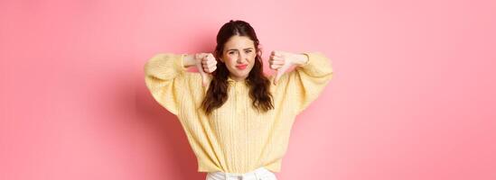 Very bad. Disappointed young woman complaining on something awful, showing dislike with thumbs down, grimacing and frowning, shaking head, give negative feedback, pink background photo