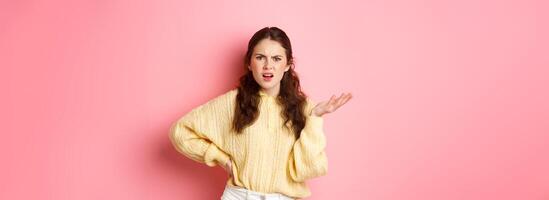 What is this, are you kidding me. Annoyed young woman complaining at something strange, staring confused, raising one hand with puzzled face, standing over pink background photo