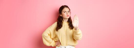 Angry and bossy young woman frowning, looking serious, showing block stop gesture, stretch out hand to say no, refuse something bad, forbid action, standing against pink background photo