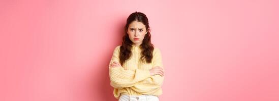 Cute silly girl feeling offended and sad, trying to comfort herself with self hug, frowning and looking at camera insulted, about to cry, standing against pink background photo