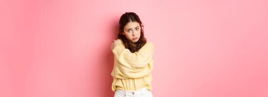 Cute silly girl making sad face and looking with puppy eyes at camera, hugging herself, standing innocent against pink background photo