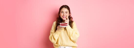 Image of excited girl thinking of eating delicious cake, biting finger from temptation and looking thoughtful at camera, think of calories in dessert, pink background photo