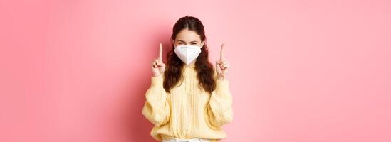 Covid, corona and social distancing concept. Thoughtful sassy girl having an interesting idea, squinting coy and smiling with eyes while wearing medical respirator, pointing fingers up at copy space photo