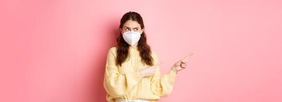 Covid, corona and social distancing concept. Confused brunette girl in medical respirator having doubts, pointing and looking aside at left copy space, standing against pink background photo