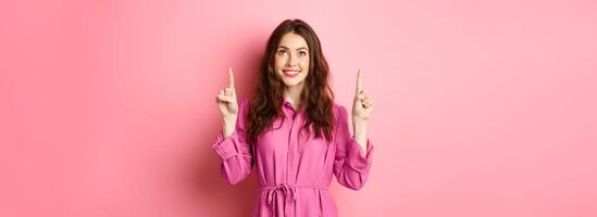 Image of beautiful happy lady in dress, pointing and looking up and smiling cheerful, staring at promo text, reading your logo, standing against pink background photo