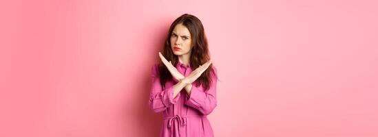 Displeased angry woman blocking offer, showing cross stop gesture, saying no and shaking head in negative reply, standing over pink background photo
