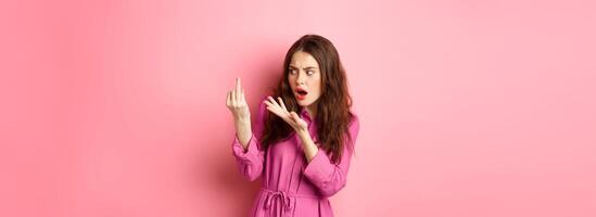 Annoyed girlfriend arguing about proposal, wants to get married, showing finger without engagement ring, standing confused against pink background photo