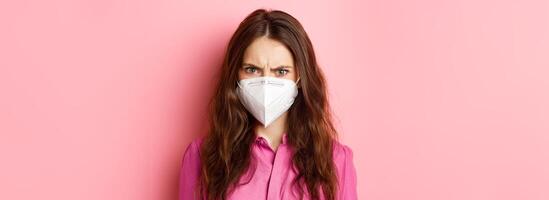 Covid-19, quarantine and social distancing concept. Close up portrait of angry female in respirator frowning, looking upset and judgemental, pink background photo