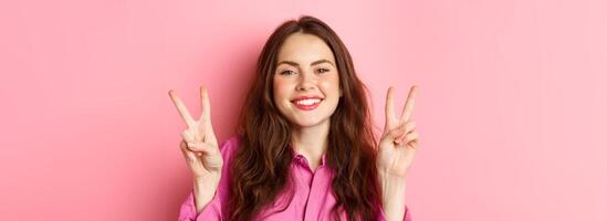 Close up of positive brunette female model showing peace signs, smiling and having fun, good vibes only, standing over pink background photo