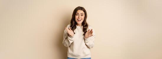Image of surprised, amazed brunette girl gasping, looking fascinated at camera, standing over beige background photo