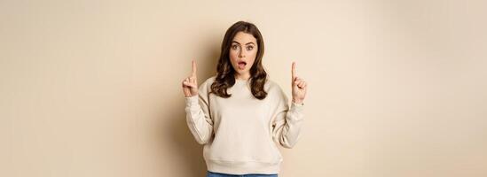 Surprised beautiful girl showing advertisement, pointing fingers on top, store sale or website link up, standing over beige background photo