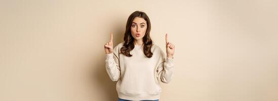Surprised beautiful girl showing advertisement, pointing fingers on top, store sale or website link up, standing over beige background photo