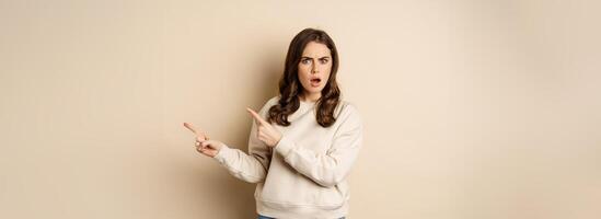 Shocked girl pointing fingers right, gasping startled, standing over beige background photo