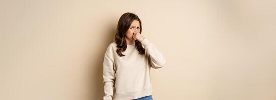 Woman expressing disgust of bad disgusting smell, close nose and grimaing from dislike, standing over beige background photo