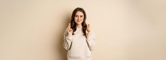 Confident woman praying, making wish with fingers crossed for good luck, standing over beige background photo
