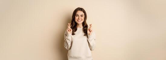 Confident woman praying, making wish with fingers crossed for good luck, standing over beige background photo