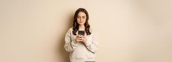 People and cellular technology. Beautiful stylish woman using mobile phone, smartphone app, smiling and looking at screen, standing over beige background photo