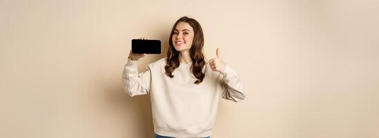 Smiling happy woman showing smartphone horizontal screen, thumbs up, recommending website, online store or app, standing over beige background photo
