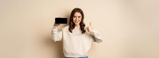 Smiling happy woman showing smartphone horizontal screen, thumbs up, recommending website, online store or app, standing over beige background photo