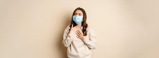 Covid-19 and health concept. Sick woman in medical face mask coughing, feeling ill with sour throat, standing over beige background photo