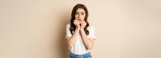 Portrait of scared woman trembling, watching smth scary and looking insecure timid, posing against beige background photo