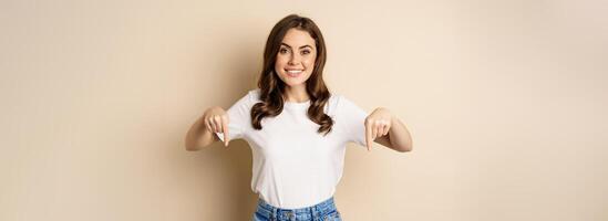 Smiling happy girl pointing fingers down and showing banner, company logo below, click link, standing over beige background photo