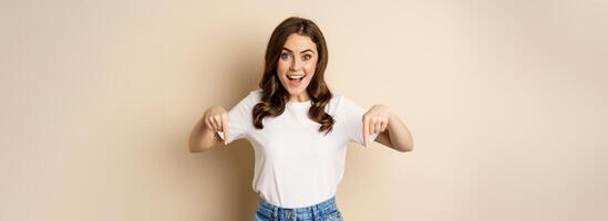 Smiling happy girl pointing fingers down and showing banner, company logo below, click link, standing over beige background photo