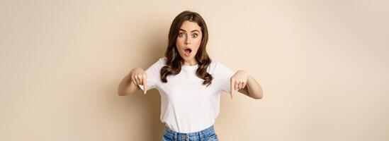 Surprised modern womn pointing fingers down, gasping amazed, showing smth awesome below, standing over beige background photo