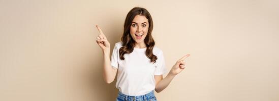 Happy authentic girl smiling, pointing fingers sideways, showing left and right banner, demonstrating promo, standing against beige background photo
