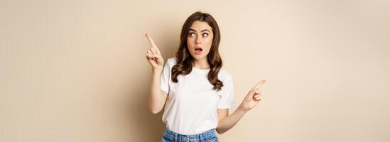 Happy authentic girl smiling, pointing fingers sideways, showing left and right banner, demonstrating promo, standing against beige background photo