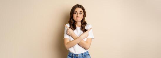 Beautiful girl choosing between two options, pointing sideways and looking clueless, standing over beige background photo