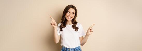 Happy authentic girl smiling, pointing fingers sideways, showing left and right banner, demonstrating promo, standing against beige background photo