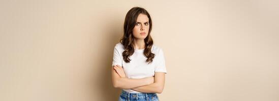 Offended angry woman cross hands on chest, frowning and sulking insulted, standing over beige background photo