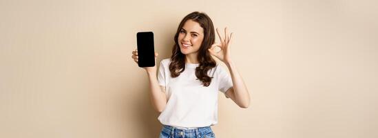 Smiling happy girl showing mobile phone screen, app on smartphone and okay, recommending online store, shopping application, beige background photo
