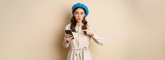 Excited girl with smartphone pointing finger at mobile screen, looking amazed, wow intrigued face expression, beige background photo